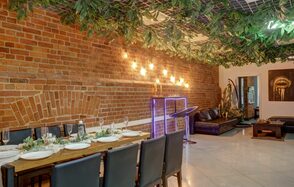 Лофт Семеновский. Пространство с кирпичными стенами и плиточным полом. Длинный деревянный стол с черными стульями, сервирован посудой и бокалами. Потолок украшен зелеными листьями и светодиодными лампами. В углу зона отдыха с кожаными диванами и столиком.