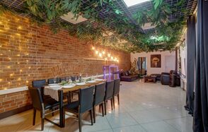 Лофт Семеновский. Праздничное пространство с кирпичной стеной и подвесными растениями на потолке. Стол со стульями сервирован для обеда. В углу мягкая мебель. На стене световые украшения. Плиточный пол, большие окна с черными шторами.