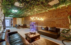 Лофт Семеновский. Лофт-пространство с кирпичными стенами и подвесным потолком, украшенным зеленью. Кожаные диваны и деревянный стол в центре. В углу стол с креслами. Светильники на стене. Яркая неоновая установка. Пол плиточный.