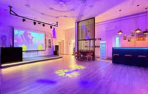 Лофт Софт. Просторное мероприятие с деревянным полом и высоким потолком, украшенным лепниной. В центре — сцена с большим экраном. Справа — барная стойка с подвесными светильниками. Красочное освещение, несколько столиков и стульев.