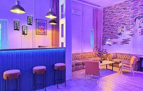 Лофт Софт. Праздничное пространство с барной стойкой, облицованной синими панелями, и четырьмя барными стульями. Левый угол оформлен в виде зоны отдыха с коричневым диваном, креслом и журнальным столиком. Стены украшены картинами и кирпичной кладкой. Освещение фиолетово-жёлтое.