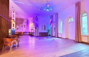 Лофт Софт. Просторное помещение с деревянным полом, цветным освещением и кирпичной стеной. По краям расположены диван и стулья, большие арочные окна с занавесками. Подвесные светильники и прожекторы обеспечивают яркую подсветку.