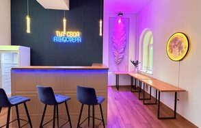 Лофт Лайф. Барная стойка с барными стульями, неоновая надпись на синей стене, подвесные светильники, декоративная панель с подсветкой, деревянные столы вдоль стены, окна с арками, деревянный пол, декоративный круг с подсветкой на стене.