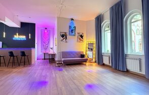 Лофт Лайф. Просторная гостиная с деревянным полом, барной стойкой с тремя черными стульями слева. На стене вывеска с подсветкой. Большое зеркало в золотой раме, диван у стены и картины с подсветкой. Окна с высокими шторами, яркое разноцветное освещение.