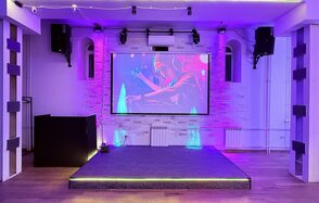 Лофт Лайф. Сцена с проектором и изображением диджея, чёрные колонки по бокам, светодиодная подсветка, паркетный пол, белые стены с декоративной кирпичной кладкой, потолочные светильники, радиаторы отопления, фиолетовое освещение.