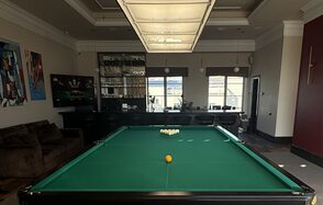 Студия Н. Бильярдный зал с большим зелёным столом в центре. Барная стойка с высокими стульями вдоль стены. Кожаный диван слева. Подвесное освещение и настенные картины. Коричневый пол, светлые стены с тёмными акцентами.