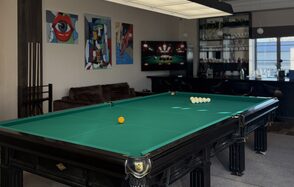 Студия Н. Бильярдная комната с столом для русской пирамиды, рядом с ним диван и телевизор, на стенах картины в ярких цветах. Стойка с напитками и бокалами, барные стулья, окно с видом на город, светлая перегородка с освещением.