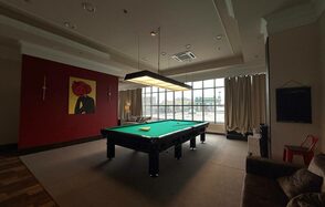 Студия Н. Бильярдная комната с зеленым столом, висячей лампой над ним, большим окном с занавесками. На стене висит картина. На полу ковровое покрытие, по бокам диван и кресло, справа красный стул. Потолочное освещение и встроенная система кондиционирования.