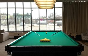 Студия Н. Бильярдный стол с зелёным сукном, пара киев, шары треугольником. Просторное помещение с большими окнами, тип штора кремового цвета справа. Потолочный светильник над столом. Слева диван и кресло с узором. Панорамный вид на городские крыши.