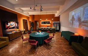 The Gallery Loft (Лофт Галерея) . Просторная комната с деревянным паркетом, круглым столом с цветами и красными стульями. Встроенный проектор, барная стойка с высокими стульями, черный холодильник. Оранжевые стены с изображением камина, потолочные прожекторы и зеркальный шар.