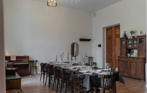 Пространство Имена. Винтажный лофт . Большая столовая с длинным деревянным столом и стульями, накрытыми черной тканью, сервировка посуды и бокалов. Слева пианино, шкаф и зеркало на стене. Деревянный пол, стены светлые. Потолочная люстра с подвесками.