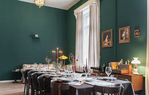 Пространство Имена. Винтажный лофт . Столовая с длинным накрытым столом на двенадцать персон, деревянные стулья, плотные белые шторы, зеленые стены, картины в рамках. На комоде лампы и декор, в центре стола свечи и цветы. Потолочная люстра, паркетный пол.