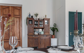 Пространство Имена. Винтажный лофт . Столовая с деревянным буфетом, на котором стоят вазы и растения. Перед буфетом накрытый стол с белыми тарелками и бокалами. Крупные деревянные двери справа закрыты, рядом зеленая дверь, рядом с буфетом комнатное растение.