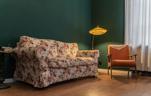 Пространство Имена. Винтажный лофт . Гостиная с темно-зелеными стенами и деревянным полом. Диван с цветочной обивкой и оранжевое кресло. Напольная лампа с тканевым абажуром, белая занавеска на окне. Маленький круглый стол рядом с диваном.