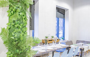 Лофт Облака. Зал Баунти на Полярной. Современное помещение с белыми стенами, окнами, столом с белыми тарелками и бокалами. Голубые стулья, светлое деревянное напольное покрытие, декоративные растения на стене и столе, черный диван у стены, естественное освещение.