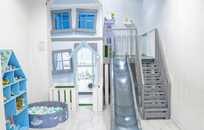 Лофт Облака. Зал Баунти на Полярной. Детская игровая зона с деревянным домиком, лестницей и горкой. Стены и пол светлые, корзина с цветными шарами. Шкаф с игрушками слева, домик окрашен в голубые и белые тона. Яркое освещение, на стене заметен текст.