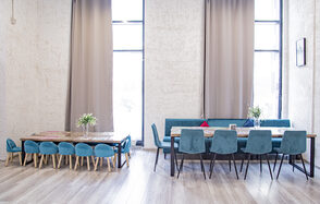 Лофт Облака. Зал Баунти на Сельскохозяйственной. Просторное помещение с двумя длинными столами и стульями с мягкой обивкой бирюзового цвета. Высокие окна с бежевыми шторами. Светлые деревянные полы. На столах цветочные композиции. Стены светлые с текстурой.