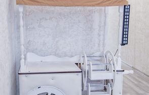 Лофт Облака. Зал Баунти на Сельскохозяйственной. Деревянная тележка с белыми и светло-коричневыми элементами, расположена у светлой стены с текстурой. Рядом светлая поверхность пола с деревянным узором. На тележке есть полки и стилизованные колеса.