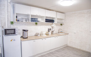 Лофт Облака. Зал Баунти на Сельскохозяйственной. Кухонное помещение с белыми шкафами и светлыми столешницами. На столе микроволновка, термопот и кухонные принадлежности. Открытые полки с посудой. Белые стены, светло-серый пол, круглые лампы на потолке. Декор из зелени.