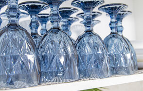 Лофт Облака. Зал Баунти на Сельскохозяйственной. Синий хрустальный бокал, перевёрнутый вверх дном, расположен на белой полке. Бокал с узором в виде геометрических форм. Поверхность полки частично покрыта текстурированной салфеткой. На заднем плане видны другие бокалы.