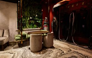 Микс Afterparty(После вечеринки). Современное помещение с зелёной стеной из растений, диваном с зелёной обивкой, круглым столом и двумя серыми креслами. Напольное покрытие из мрамора с волнистыми узорами. Красные стены, настенное освещение, справа часть гардеробной стойки.