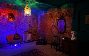 Лофт «Рич» (RICH). Интерьер с кирпичными стенами; потолочное освещение в виде люстры; мягкое кресло с гитарой; зеркало в резной раме; статуя на тумбе; ковёр на полу; разноцветная подсветка; картины на стене; табурет и радиатор вдоль стены; тёмные и теплые цвета.