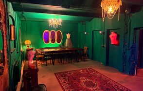 Лофт «Рич» (RICH). Комната с зелеными стенами и низким потолком. Длинный деревянный стол окружен стульями, слева бюст, на столе лампа с абажуром. Потолочные и настенные светильники. На полу яркий ковер с узорами. На стене три зеркала и розовый торс.