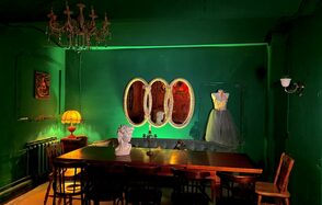 Лофт «Рич» (RICH). Зал с зелеными стенами, длинный деревянный стол с стульями, бюст на столе. Слева торшер с желтым абажуром, на стене картина. Справа платье на манекене. Тройное овальное зеркало на стене. Подвесная люстра.