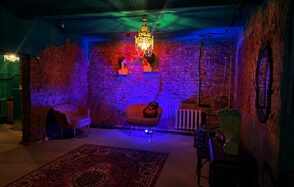 Лофт «Рич» (RICH). Кирпичные стены, два кожаных кресла, кресло с гитарой, потолочная люстра, разноцветное освещение, радиатор, ковер с узором, зеркало на стене, бюст на столике, бетонный пол.