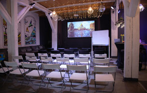 Сатурн Лофт. Современное лофт-пространство с деревянными балками и потолком, оборудованное белыми складными стульями, проектором с изображением города, экраном, флипчартом. На стенах картины, гирлянды лампочек, зона с диваном, черные барные столы.