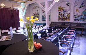 Сатурн Лофт. Пространство с рядами черных и белых стульев, черные круглые столы, белые деревянные балки, яркие картины на стенах, фиолетовая шторка, ноутбук, ваза с желтыми цветами, светильники в клетках, два граната.