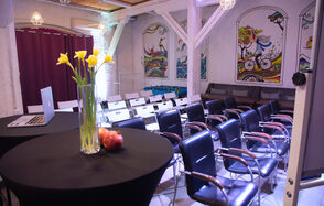 Сатурн Лофт. Пространство для мероприятий с рядами стульев и обеденными столами. Стулья черные с деревянными подлокотниками; столы покрыты черной тканью, на одном из которых ваза с желтыми цветами и яблоки. Белые стены с яркими настенными рисунками. Потолок деревянный. Освещение теплое.