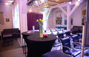 Сатурн Лофт. Лофт-пространство с белыми кирпичными стенами, деревянным потолком и балками. Ряд белых стульев, черные столы с вазой и фруктами. Настенная роспись, фиолетовые шторы. Потолочные светильники с лампочками. Черные кресла и диван.