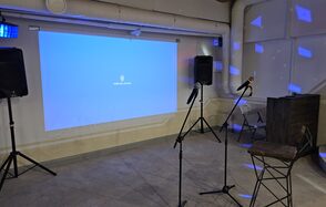 Караоке-лофт с бильярдом. Клубное пространство с проекционным экраном и двумя микрофонами на стойках. Два крупных динамика по бокам. Стул с высокой спинкой рядом с микрофонами. Серый пол, стены светлые. Подвесные трубы и вентиляция вверху. Синие световые эффекты на стенах.