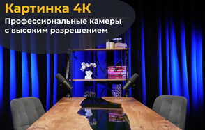 Лофт Восторг. Современное пространство с деревянным столом и серыми стульями. На столе черные микрофоны. Позади полка с книгами и декором, включая цветы. Задник оформлен темными шторами с синим освещением. Текст «Картинка 4К: Профессиональные камеры с высоким разрешением».