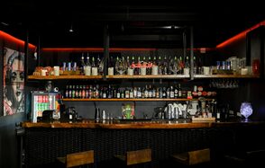Аутентичный просторный лофт. Барная стойка с деревянной столешницей и тремя стульями, полки с бутылками и предметами, фужеры подвешены к стойке. Освещение включено в потолке, неоновые красные акценты. Темные стены с графическим изображением.