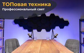 Лофт Позитив. Современное пространство с деревянным столом и двумя серыми креслами. На столе стоят микрофоны и стаканы с водой. На стене шестиугольные черные панели. Освещение фиолетовое. Слева стеллаж с книгами. Надпись: «ТОПовая техника. Профессиональный свет».