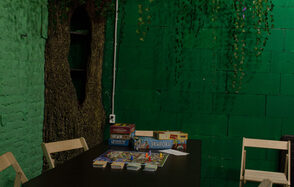 Хендмейдщики. Лофт в лесной тематике. Зал с зелёными кирпичными стенами, декорацией в виде ствола дерева и висящей листвой. Чёрный стол с настольными играми и карточками. Деревянные стулья. Освещение уютное и приглушённое, розетка на стене.