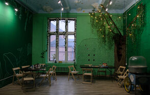 Хендмейдщики. Лофт в лесной тематике. Зеленое помещение с высоким потолком и декоративными элементами. Стены оформлены в виде деревьев с гирляндами. Несколько складных столов и деревянных стульев. Окно с белыми рамами и решеткой. На полу деревянные панели. Освещение точечными светильниками.