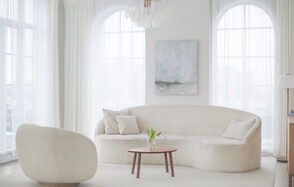 Mimika(Мимика). Просторная фотостудия. Светлая гостиная с большими арочными окнами и белыми шторами. Овальный диван и кресло в светлых тонах, небольшой деревянный кофейный столик с вазой и цветами. На стене картина. Люстра с прозрачными элементами. Светлые стены и ковер.