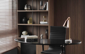 Mimika(Мимика). Просторная фотостудия. Офисное пространство с овальным столом из темного дерева, черным кожаным креслом, лампой с медным абажуром. Фоном служит деревянный стеллаж с книгами и декором. Слева жалюзи на окне, пропускающие свет. Цвета: коричневый, черный, медный.