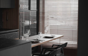 Mimika(Мимика). Просторная фотостудия. Современное помещение с деревянным столом у окна с жалюзи. На столе сервировка для двух человек: тарелки, бокалы, приборы. Два черных стула, темные деревянные элементы, светлая и серая гамма интерьера.