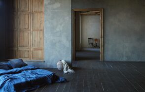Зал Flo. Фотостудия Мимика. Пространство с деревянными полами и серыми стенами, левая стена состоит из деревянных панелей. На полу синий матрас с подушками, рядом глиняный горшок с тканью. Через дверной проём видна комната с деревянным стулом и столом.