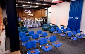 MPSellers(МПСеллерс). Просторный зал для мероприятий. Пространство зала с рядами голубых стульев и мягкой мебелью у кирпичной стены. Барная стойка с высокими стульями, задник из белого кирпича. Синий занавес с надписями. Металлические балки на потолке, ковровое покрытие. Надпись на стене: "MPG..."