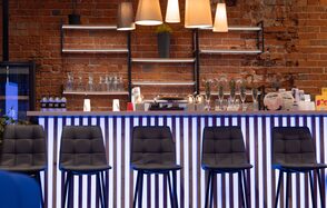 MPSellers(МПСеллерс). Просторный зал для мероприятий. Барная стойка с высокими черными стульями, кирпичная стена, полки с бутылками и бокалами, несколько ламп с абажурами. Бело-голубая подсветка стойки, на полке растение в горшке.