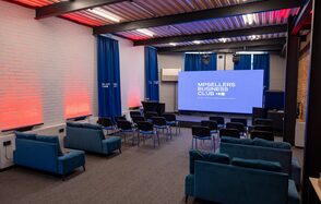 MPSellers(МПСеллерс). Просторный зал для мероприятий. Аудитория с проектором и экраном, расстановкой стульев в нескольких рядах. Несколько диванов сзади, синие и красные светодиодные светильники на кирпичных стенах, синие театральные шторы, металлический потолок, серый ковролин, кондиционер на стене.