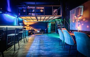 Лофт ZUM(Зум). Просторное помещение с диванами и креслами, барной стойкой и высокими стульями. На стенах неоновая подсветка и проектор с изображением микрофона. Лестница ведет на второй уровень. Напольное покрытие из плитки, освещение мягкое, разноцветное.