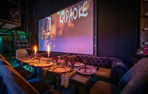 Лофт ZUM(Зум). Караоке-зал с приглушенным светом, длинный деревянный стол с сервировкой для вечеринки, бархатные диваны и стулья. На стене большой экран с изображением микрофона и надписью "Караоке". Подсвеченные лестницы и светодиодные огни.