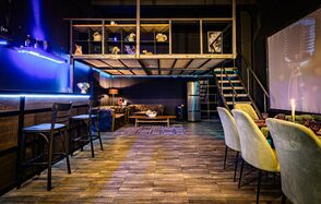 Лофт ZUM(Зум). Лофт-пространство с открытым вторым уровнем, лестницей, барной стойкой с высокими стульями, диванами, деревянным столом. Деревянный пол, мягкие стулья, декоративные огни, металлический каркас. Тёмные стены, яркое освещение, металлический холодильник.