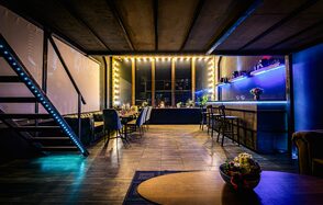 Лофт ZUM(Зум). Лофт-пространство с лестницей, вдоль стены большой стол с черными стульями, освещение от гирлянд и светодиодов. Барная стойка с высокими стульями, пол деревянный, потолок из темных панелей. Синие и золотые тона, присутствуют цветы.